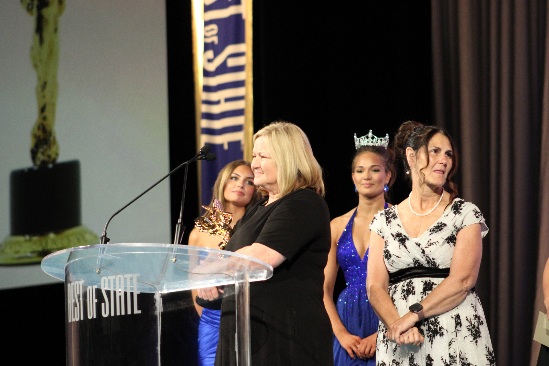 An honoree accepts the BOSS® at the Best of State podium with Miss Utah royalty looking on.